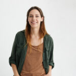 Pleased pretty woman with dyed hair, dark eyes and healthy skin dressed in brown T-shirt, green jacket holding hands in pockets smiling while posing against white concrete wall. People and lifestyle